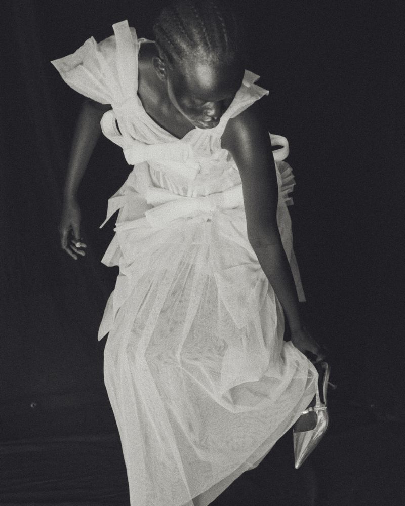 A woman in a white tulle dress is photographed against a dark background, holding a silver high-heeled shoe in a fashion editorial style.