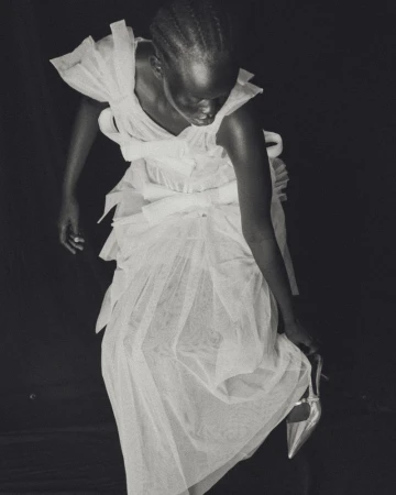 A woman in a white tulle dress is photographed against a dark background, holding a silver high-heeled shoe in a fashion editorial style.