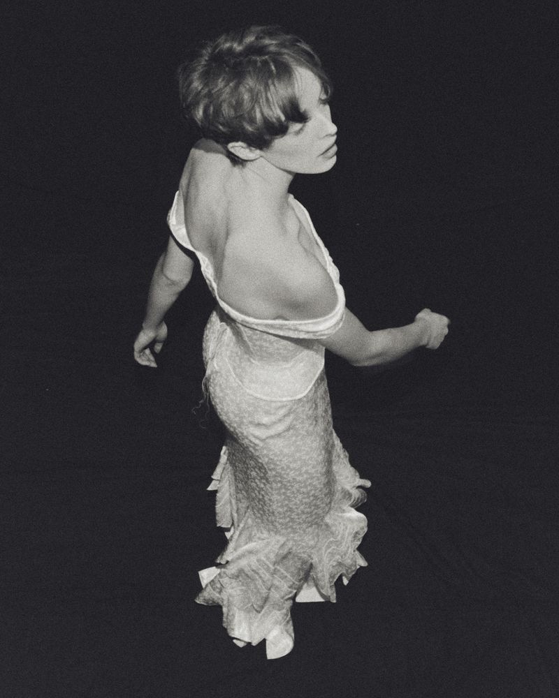 Monochrome portrait of a slender woman with short hair wearing an open-back dress, with a black backdrop in a studio setting.