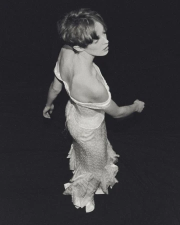 Monochrome portrait of a slender woman with short hair wearing an open-back dress, with a black backdrop in a studio setting.