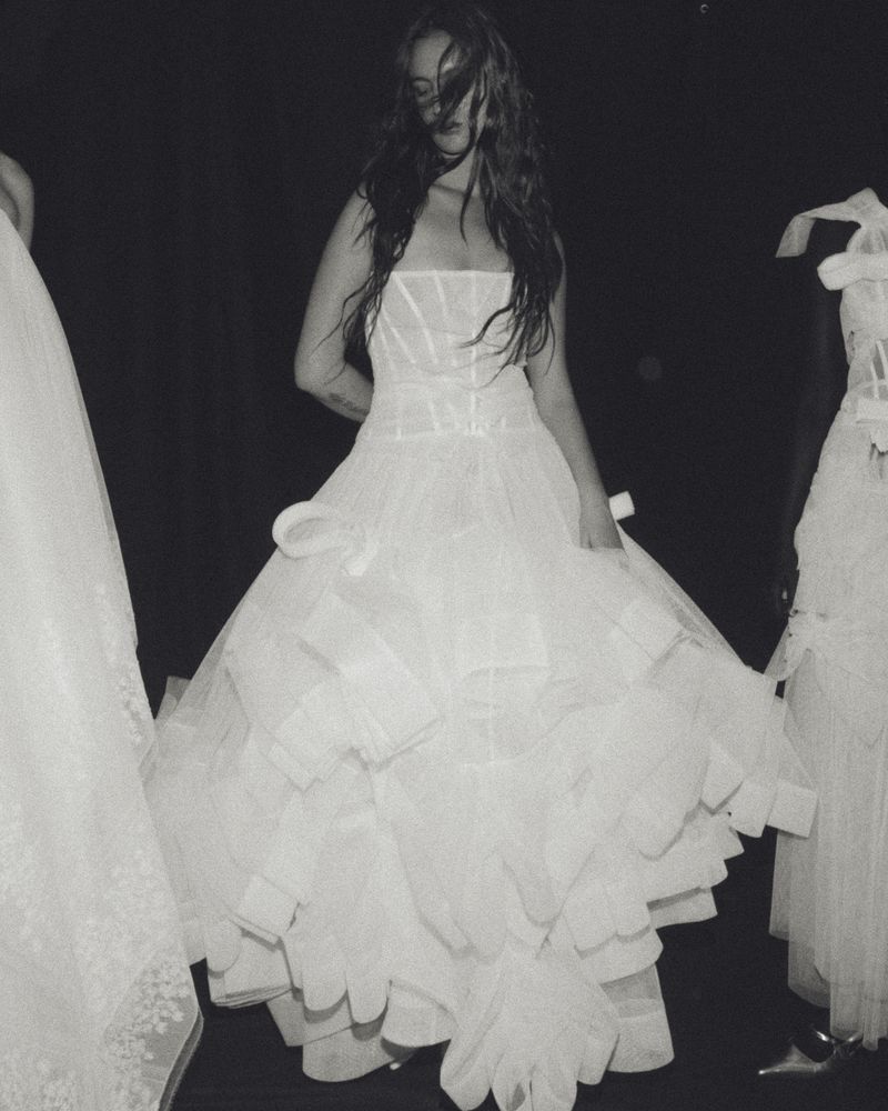 Monochrome image of a woman in a white strapless gown with a ruffled skirt, posing between two other similarly-dressed women in gowns.
