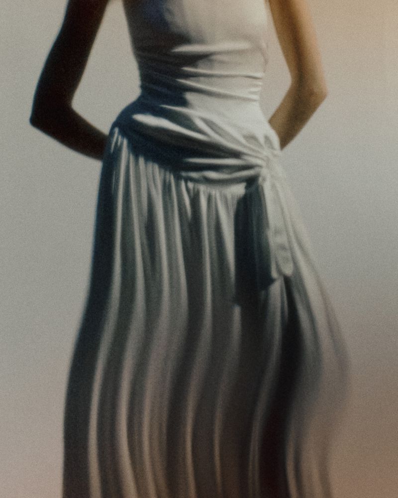 Model wearing a gathered white draped bridal gown with pleated skirt details against a softly lit studio background.