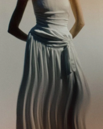 Model wearing a gathered white draped bridal gown with pleated skirt details against a softly lit studio background.