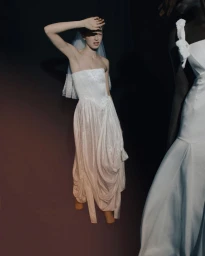 Model wearing a strapless draped white bridal gown and tulle veil, shielding her face with her hand against a dark backdrop.