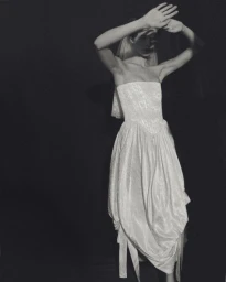 Model wearing a strapless draped white bridal gown and veil raising her hands to her forehead against a black background.
