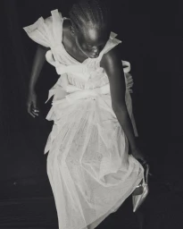 Black female model in a white tiered tulle bridal gown adjusting her pointed silver heel against a black backdrop.