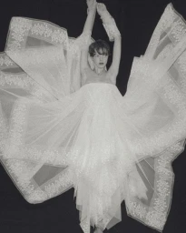 Model posing with arms raised in an expansive white tulle bridal dress with lace embroidery against a solid black background.