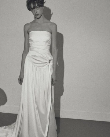 Model wearing a strapless white draped wedding gown against a plain studio wall casting a sharp shadow.
