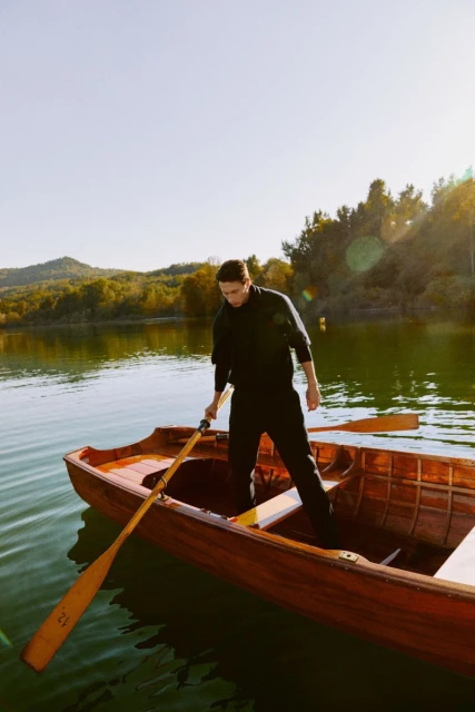 A man in dark clothing stands in a wooden rowboat with oars on a sunny lake, with green trees and distant mountains.