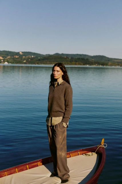 A young woman in a brown sweater and pants stands in a wooden boat on a lake with trees and buildings visible on the distant shore.