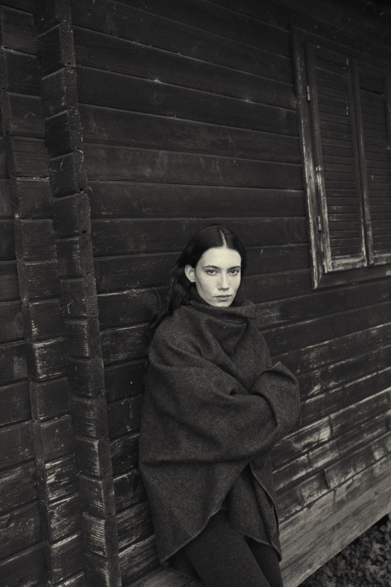 Monochrome portrait of a woman with long hair wrapped in a shawl, leaning against a dark wooden building with a window.