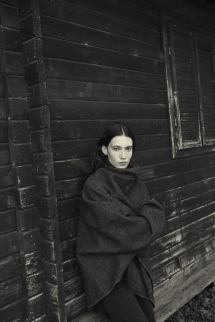 Monochrome portrait of a woman with long hair wrapped in a shawl, leaning against a dark wooden building with a window.
