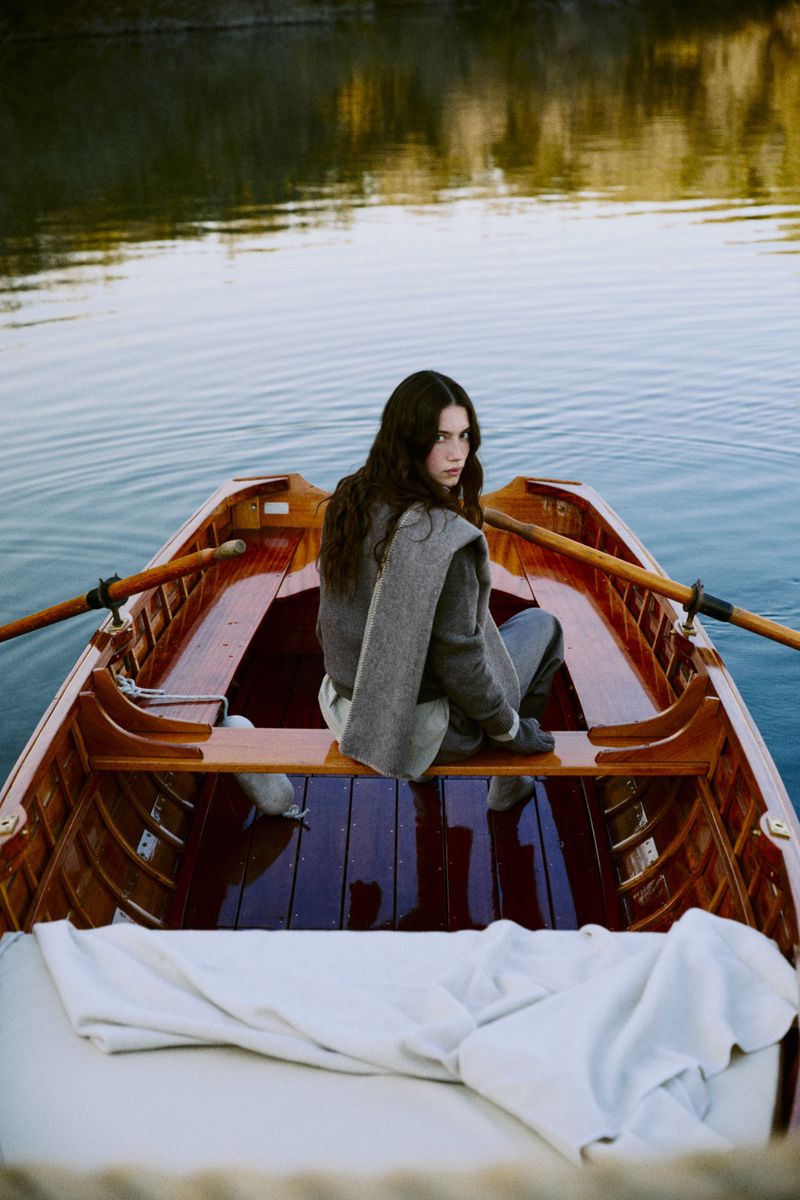 Young woman with long dark hair sits in a wooden rowboat on a calm lake with oars, gazing over her shoulder toward the camera.