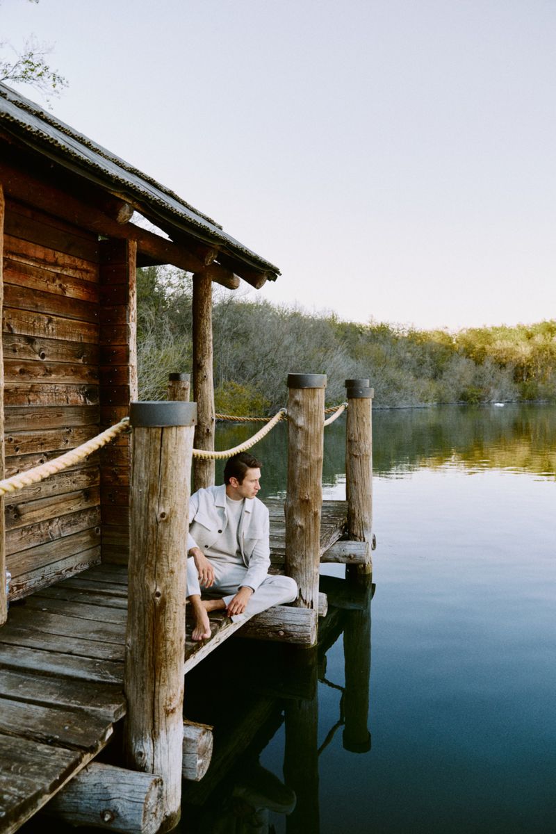 Adult male in casual clothing sits barefoot on a wooden dock at a lake house near a calm lake surrounded by trees in natural light.