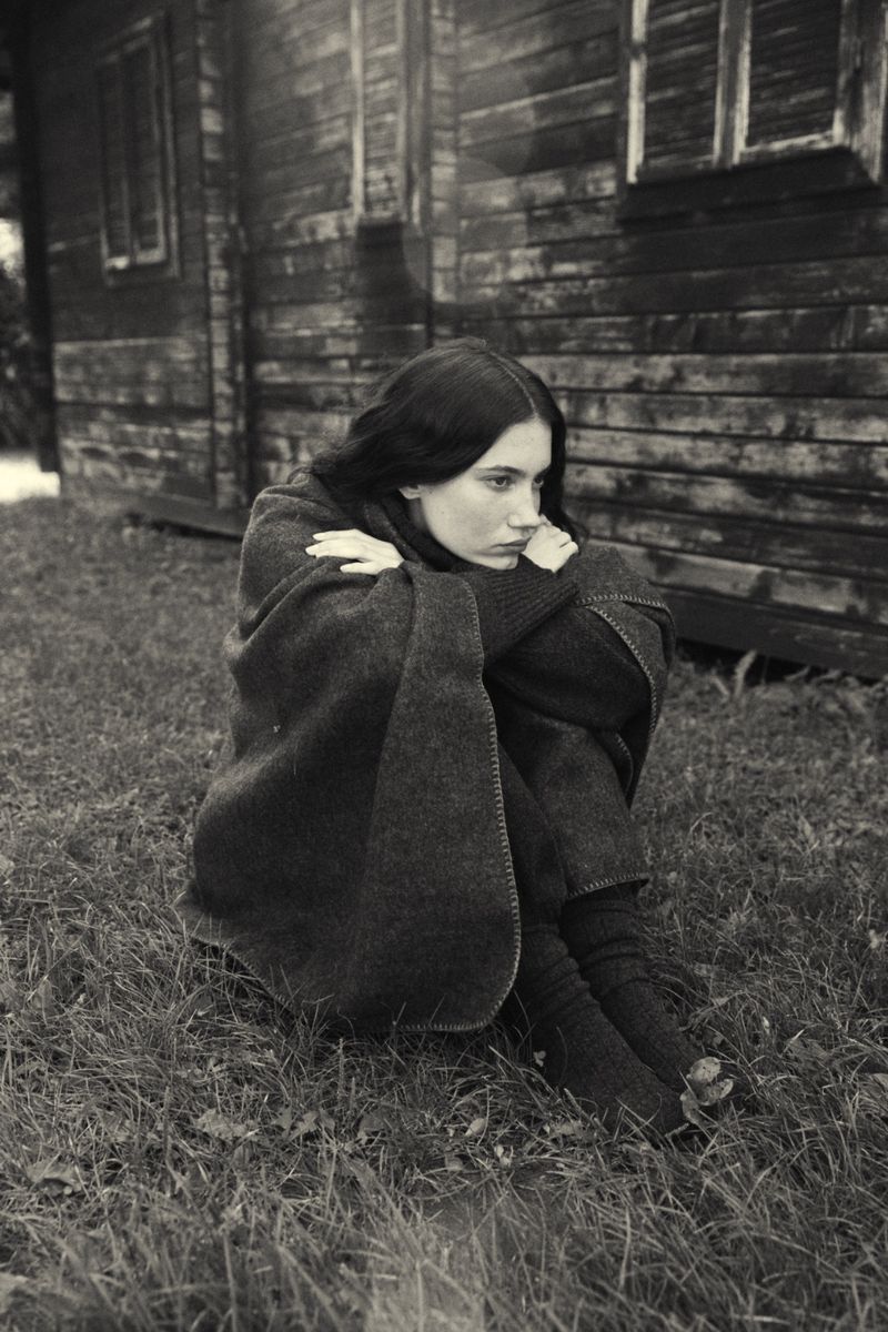 Young woman wrapped in a blanket sits outdoors near a weathered wooden building in a black and white, portrait-oriented shot.