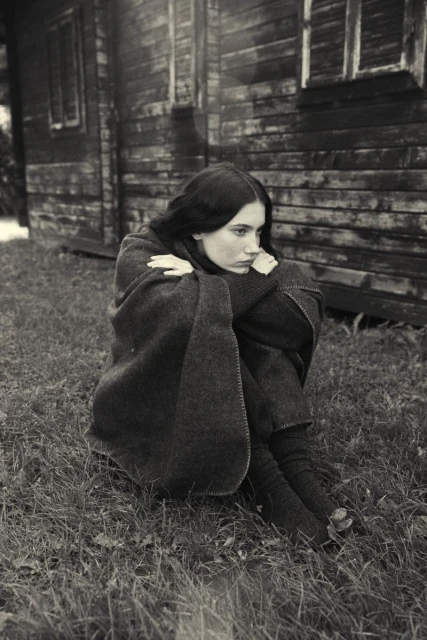 Young woman wrapped in a blanket sits outdoors near a weathered wooden building in a black and white, portrait-oriented shot.