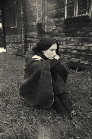 Young woman wrapped in a blanket sits outdoors near a weathered wooden building in a black and white, portrait-oriented shot.