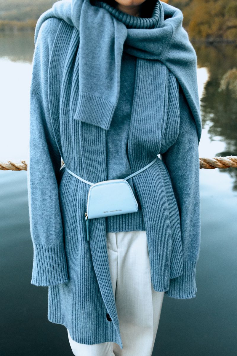Woman wears blue knitwear and white pants near lake. A small blue purse is attached to the waist with a thin strap, creating a fashionable look.