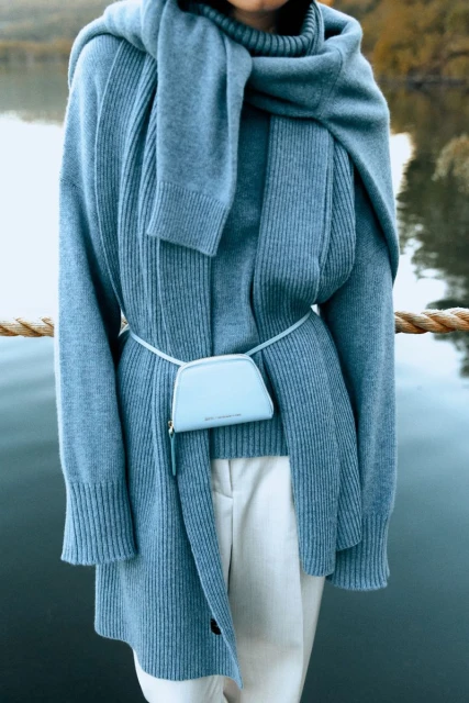 Woman wears blue knitwear and white pants near lake. A small blue purse is attached to the waist with a thin strap, creating a fashionable look.