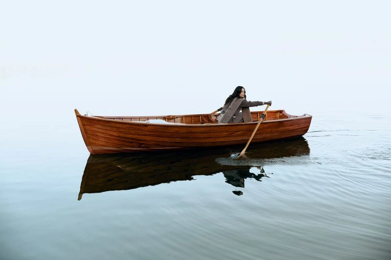 A woman in a business suit rows a wooden rowboat across the water under a bright sky, creating a sense of serenity and solitude.