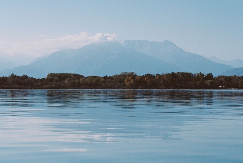 Calm lake water with a tree-lined shore and hazy blue mountains under a pale sky.