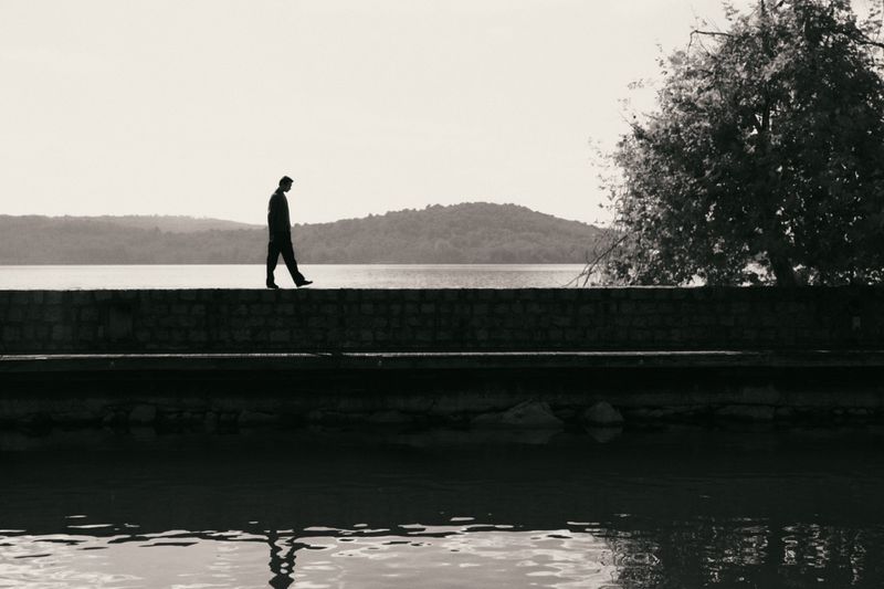 Monochromatic shot of a lone adult walking on a stone wall near a lake, mountains, and tree under an overcast sky, creating a serene scene.
