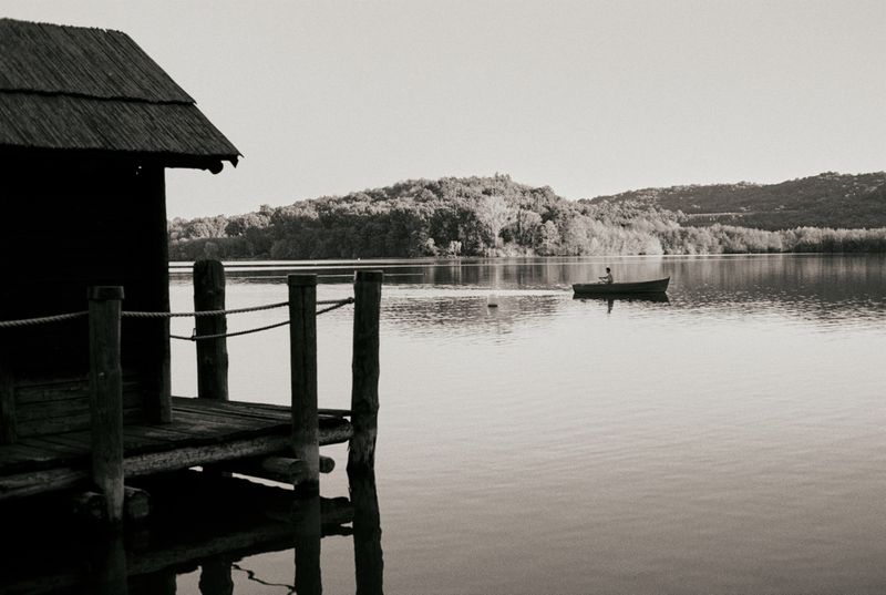 Monochrome lake scene with wooden dock, still water reflecting trees, and distant rower in boat on a calm day.