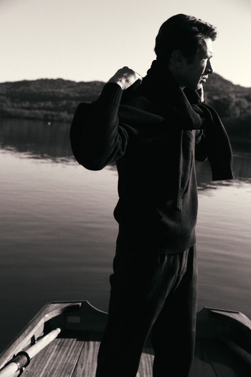 Monochrome portrait of a man in a sweater standing in a wooden boat on a lake with mountains in the distance at daytime.