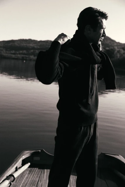 Monochrome portrait of a man in a sweater standing in a wooden boat on a lake with mountains in the distance at daytime.