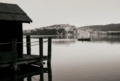 Monochrome view of a calm lake with a wooden thatched hut on a pier in the foreground and a person rowing a small boat in the distance.