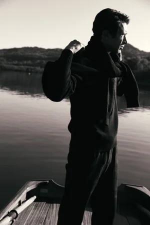 Model Victor Gorincioi standing on a wooden rowboat wearing a black knit sweater in the Artknit Studios AW25 campaign.