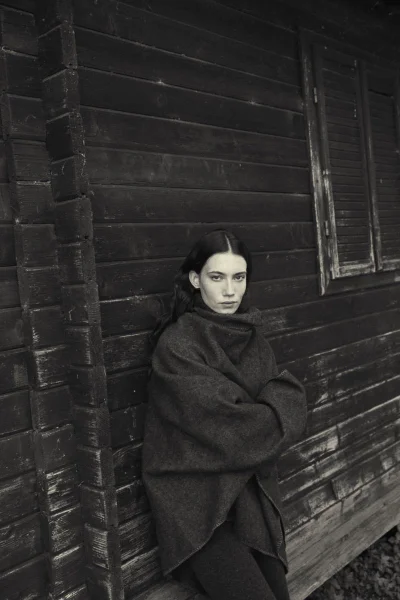 Model in a dark oversized knit sweater leaning against a rustic dark wooden wall for the Artknit Studios AW25 campaign.