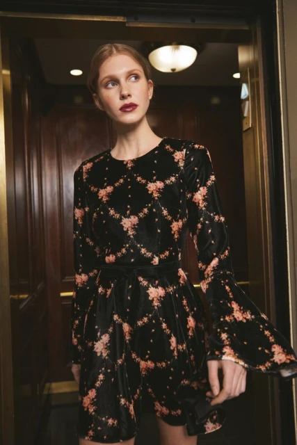 Elegant woman in a black velvet floral-print mini dress posing in a hotel elevator with wood paneling and soft, ambient lighting.