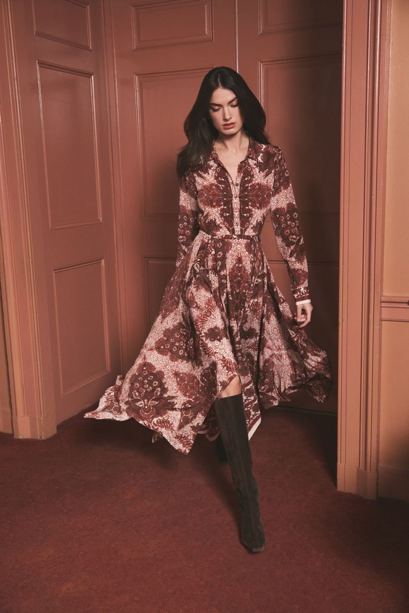 A brunette woman models a brown and white patterned dress and black knee-high boots in a room with brown walls and floors.