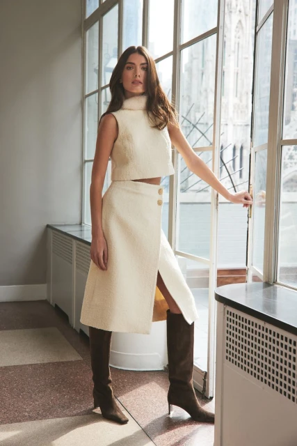 A young woman in a cream-colored, sleeveless crop top and skirt stands by a window with a cathedral in the background.