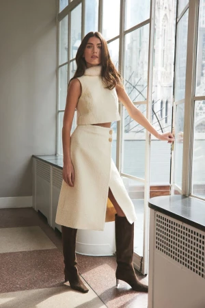 A young woman in a cream-colored, sleeveless crop top and skirt stands by a window with a cathedral in the background.