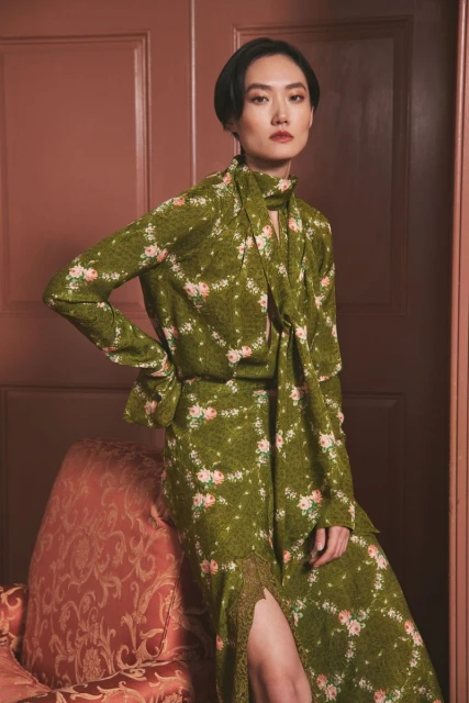 Adult female wearing a floral dress with a neck tie poses in front of a door with an upholstered arm chair in a portrait shot.