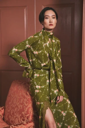 Adult female wearing a floral dress with a neck tie poses in front of a door with an upholstered arm chair in a portrait shot.