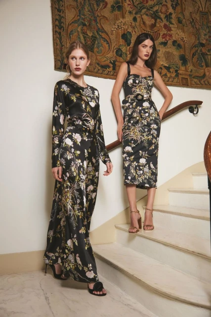 Two fashion models pose on marble stairs in floral-print dresses. One has long sleeves; the other is sleeveless. A tapestry hangs behind them.