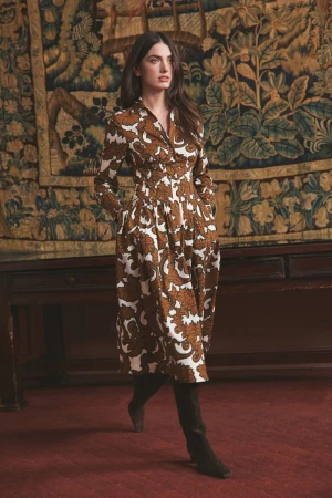 Model in a brown and white patterned midi dress with dark knee-high boots posing before an ornate woven tapestry.