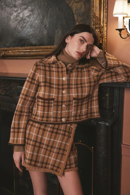 A woman poses in a tan plaid jacket and skirt with dark brown trim, leaning against a fireplace, with a vintage-style painting in the background.