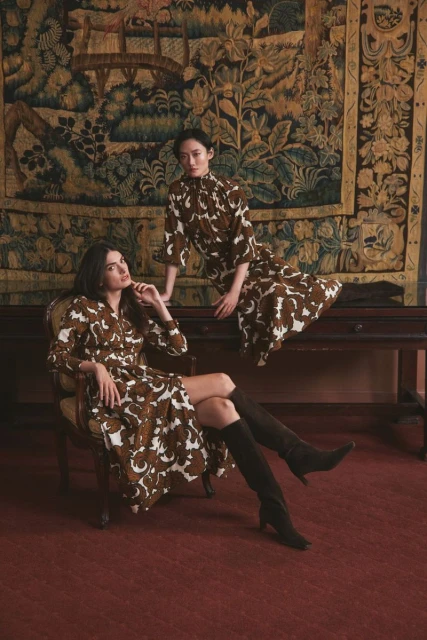 Two women pose in brown patterned dresses in an indoor setting with an antique rug and wooden table for a fashion editorial.