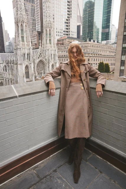 Adult female in a patterned coat and skirt poses on a rooftop with a cityscape and church in the background on an overcast day.