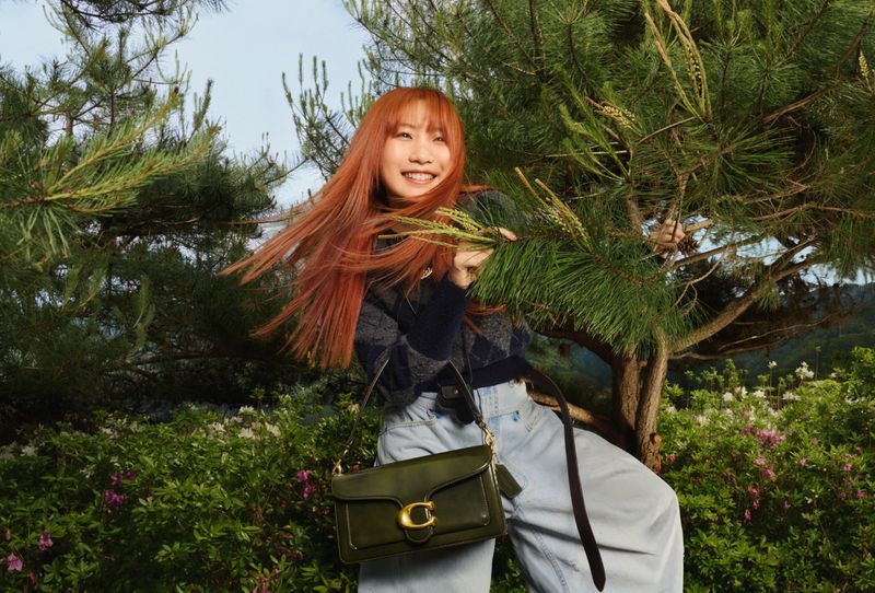 Smiling red-haired woman with a green Coach purse poses in a sunlit outdoor setting amid trees and foliage. Fashion and lifestyle image.