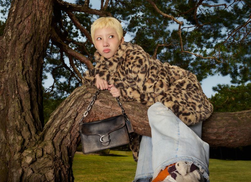 Young woman with blonde hair in a leopard print coat sits in a tree, holding a black leather bag with chain strap, outdoors.
