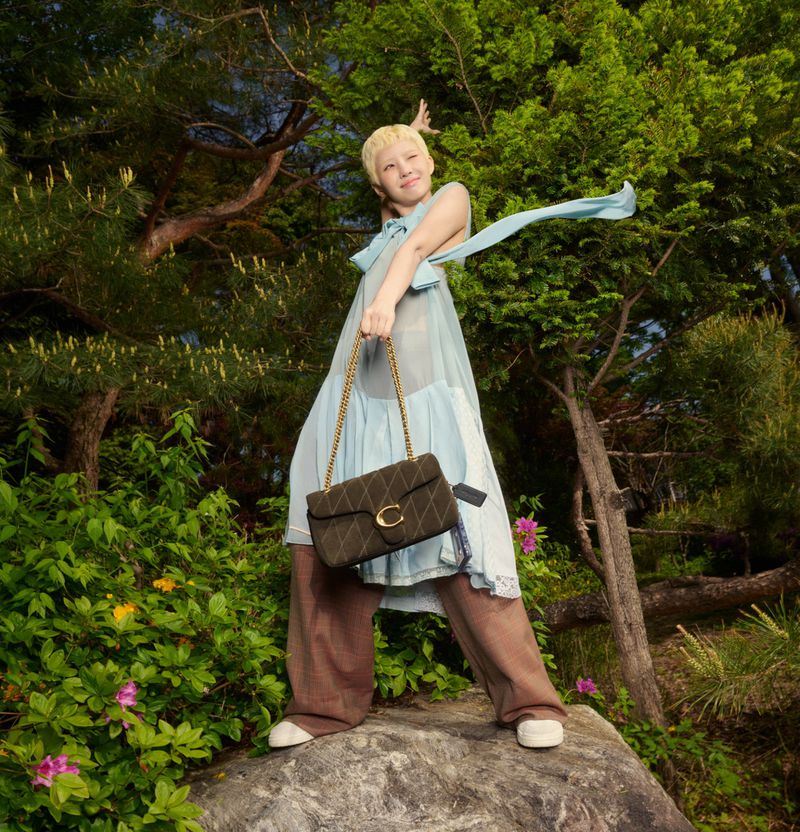 Woman standing on rock in sheer dress, holding a Coach purse with gold chain, with lush green trees and shrubbery in the background.