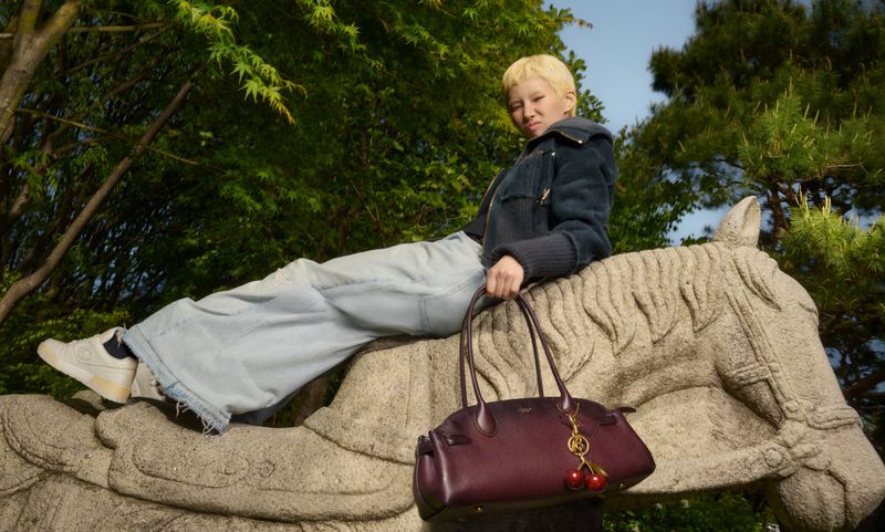 Blonde-haired woman sits atop a stone horse statue in a park, wearing wide-leg jeans, a jacket, and sneakers, and holding a red handbag.