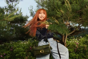 Woman with red hair smiling from a pine tree while holding an olive green Coach shoulder bag in wide-leg jeans.