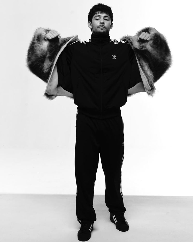 Monochrome portrait of a young adult male posing in black Adidas tracksuit with white stripes and fur jacket in a photo studio setting.