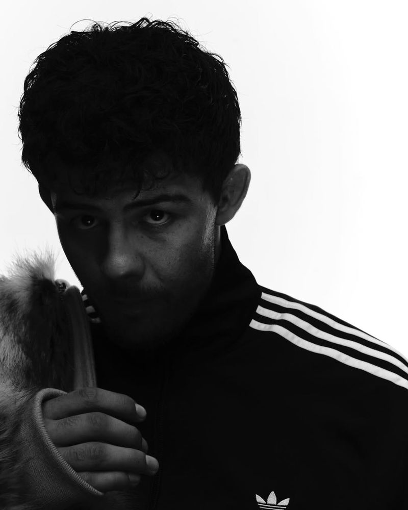 Monochrome portrait of a young adult male wearing an Adidas tracksuit jacket and holding a tan-colored furry object against a bright background.